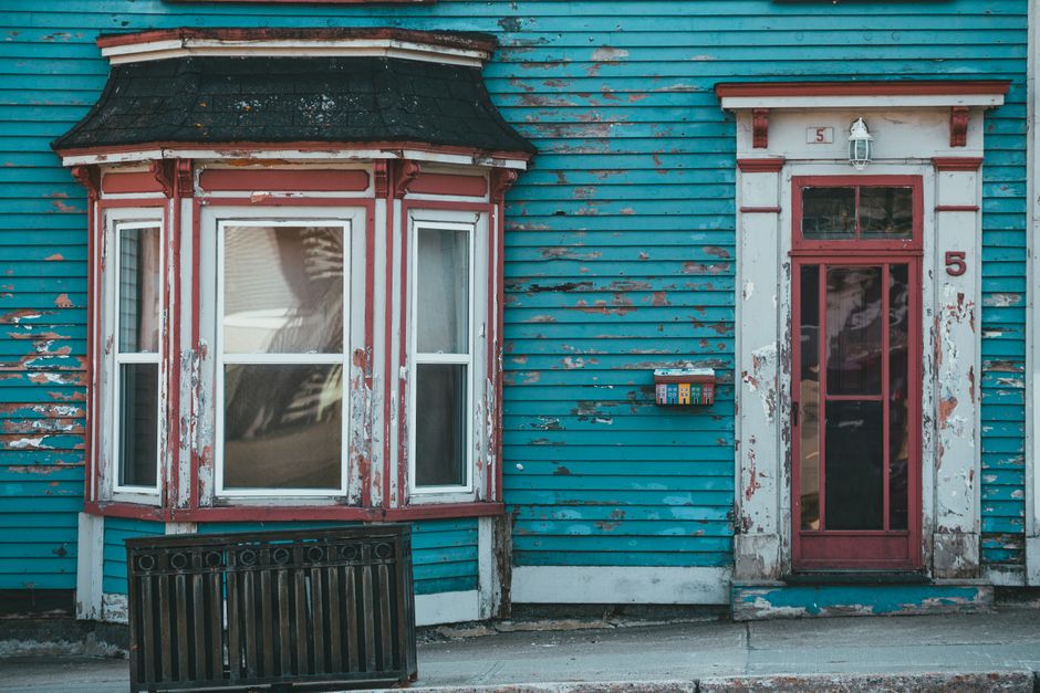 Shabby blue wall of aged residential building with bow window and door near pavement in daylight